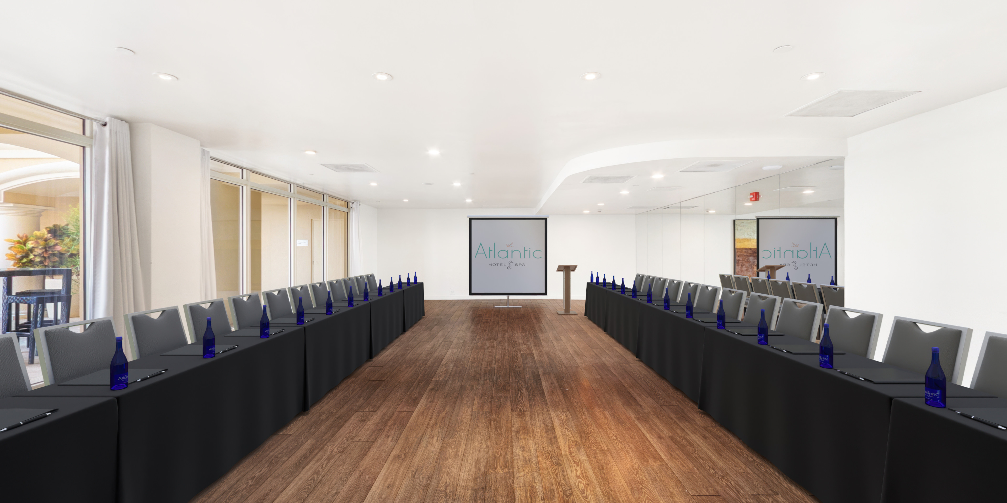 The Atlantic oceanview conference room in U-shape setup — dark tables, silver chairs, blue water bottles, hardwood floors and two projection screens displaying the Atlantic Hotel & Spa logo
