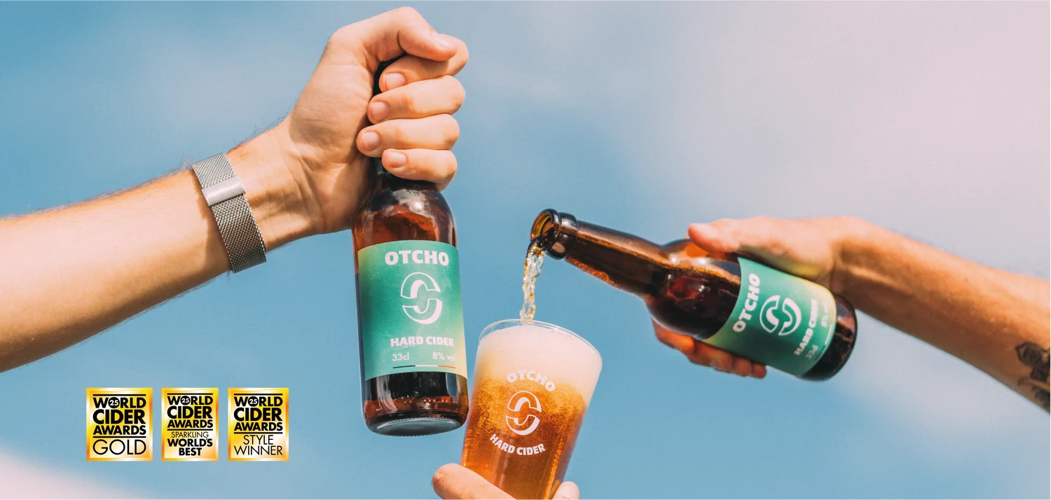 Deux bouteilles d'Otcho Cider trinquent et le cidre est versé dans un verre, devant trois médailles d'or aux World Cider Awards