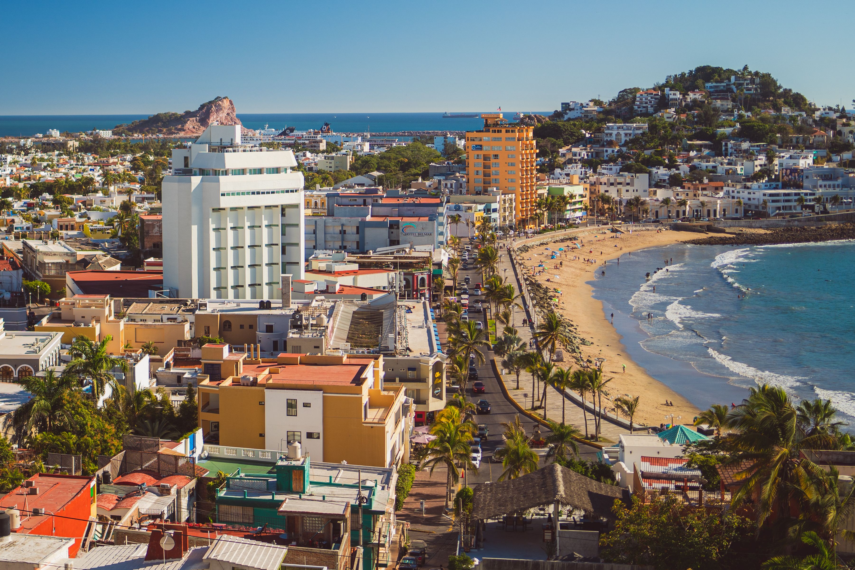 Aerial panoramic view of Mazatlán's beachfront, malecón, and iconic white resort buildings against a clear blue Pacific horizon