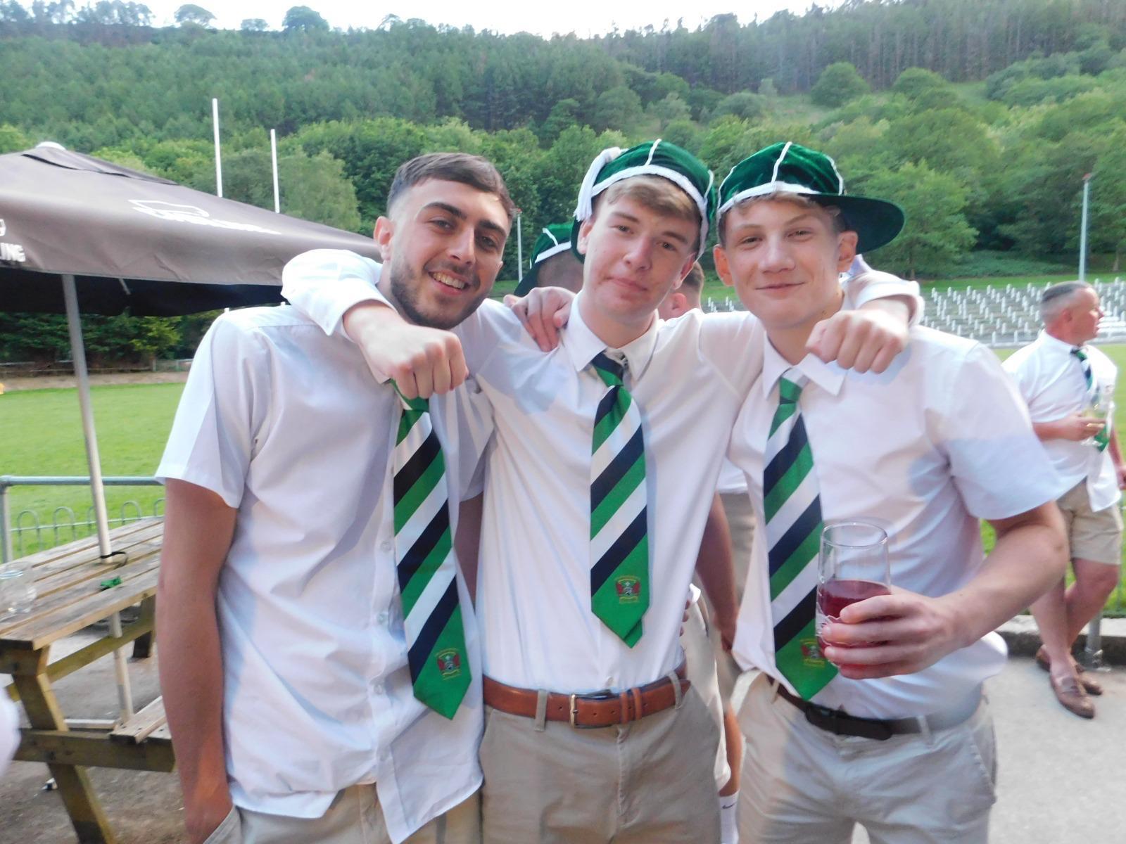 Three young men in white shirts with arms around each other, all wearing green and navy striped club ties with crest