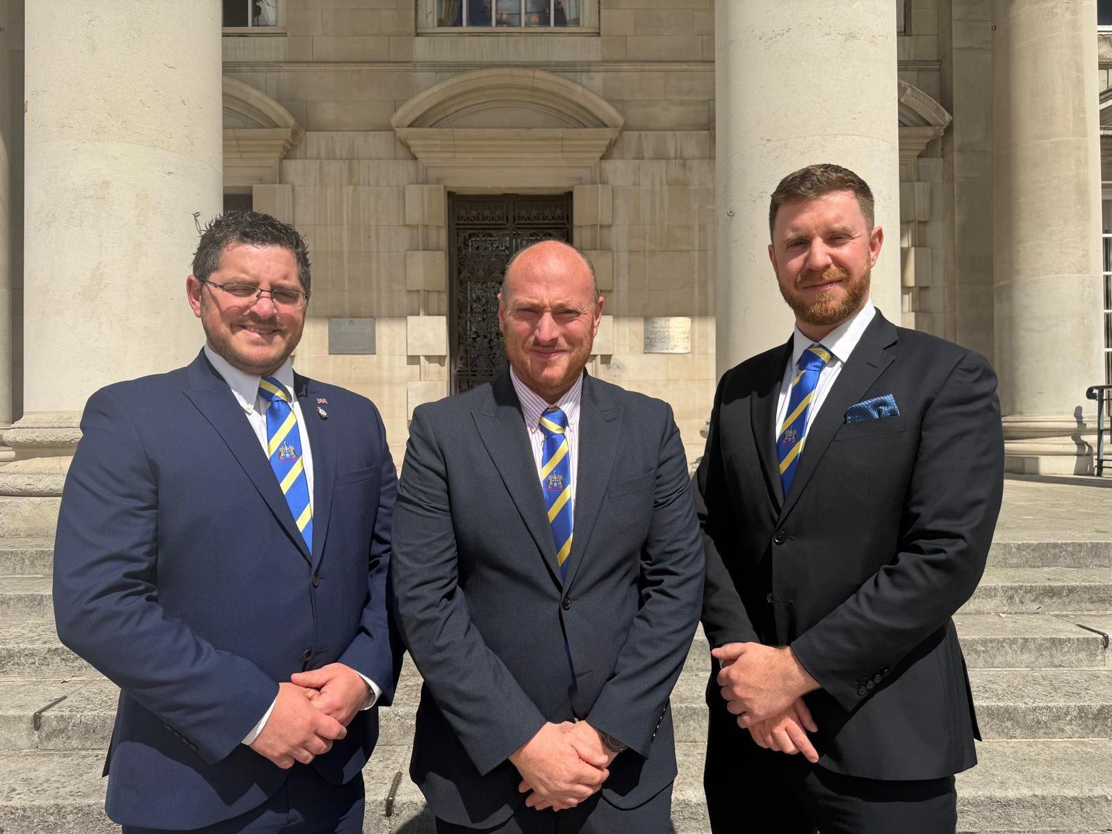 Three men in formal suits wearing royal blue and yellow striped club ties with crest outside a stone building