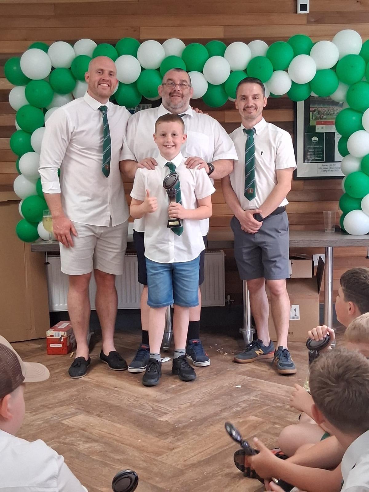 Three men in white shirts and shorts with a young boy, all wearing green striped club ties in front of a green and white balloon arch