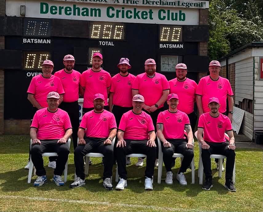 Dereham Cricket Club team in matching pink matchday shirts and caps posing in front of their scoreboard