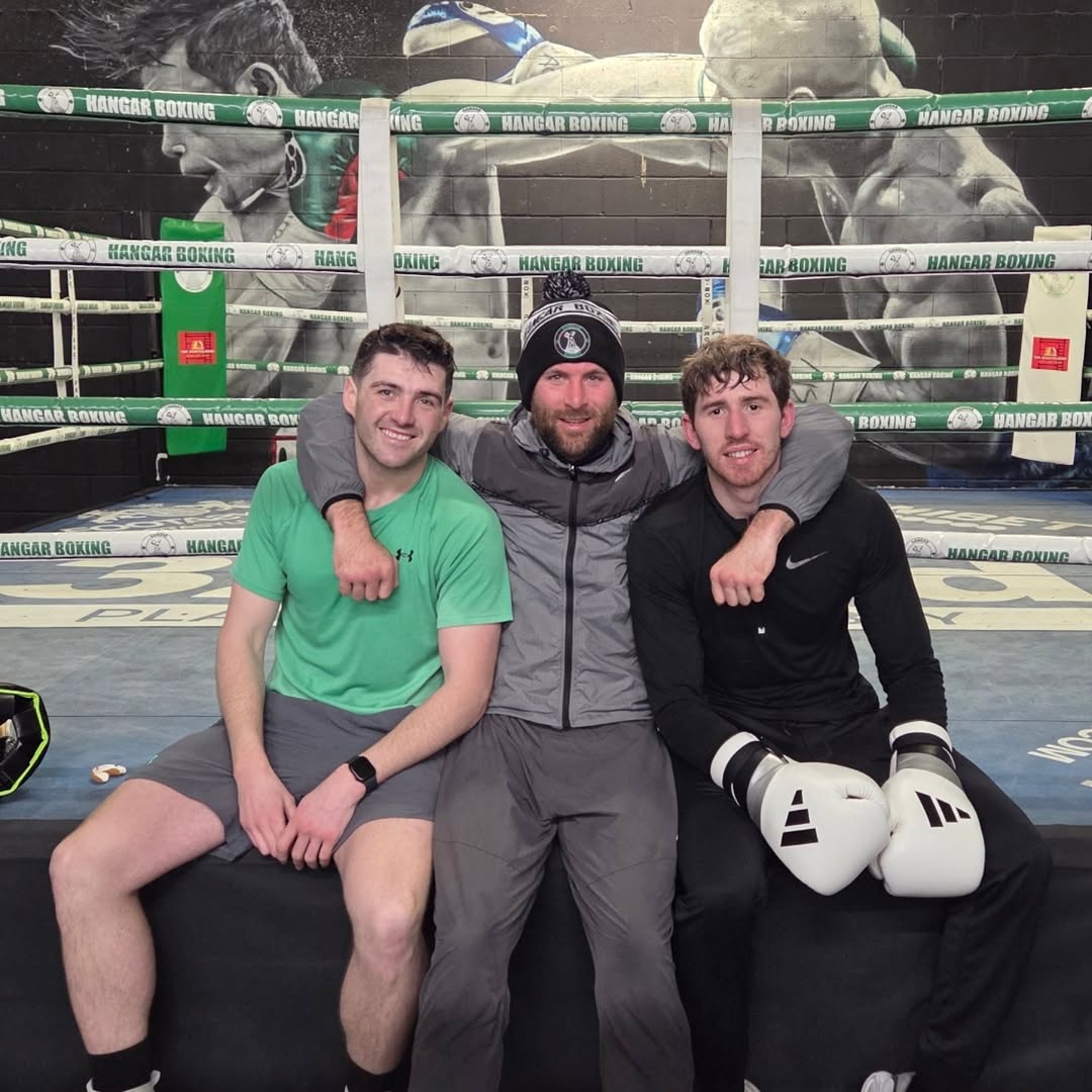 Three Hangar Boxing boxers posing together on the ring apron in front of the green and white Hangar Boxing ring ropes