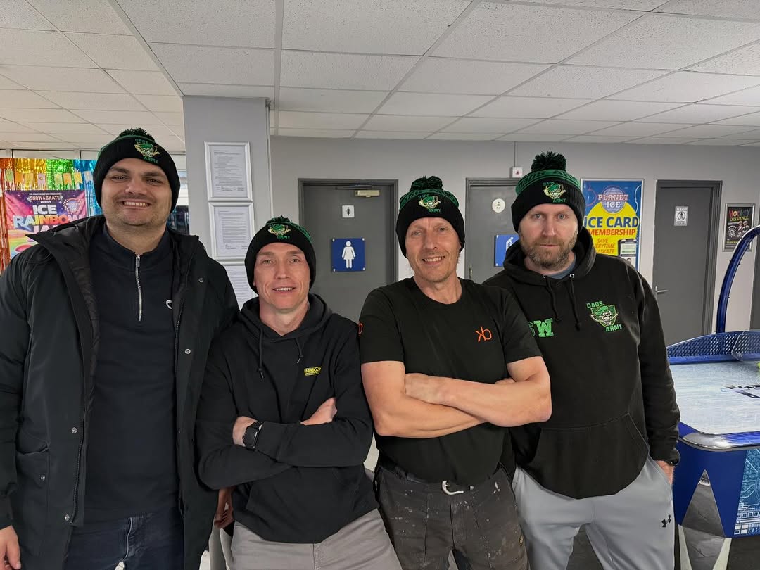 Four Dads on Ice members wearing green and black custom bobble hats in an ice rink