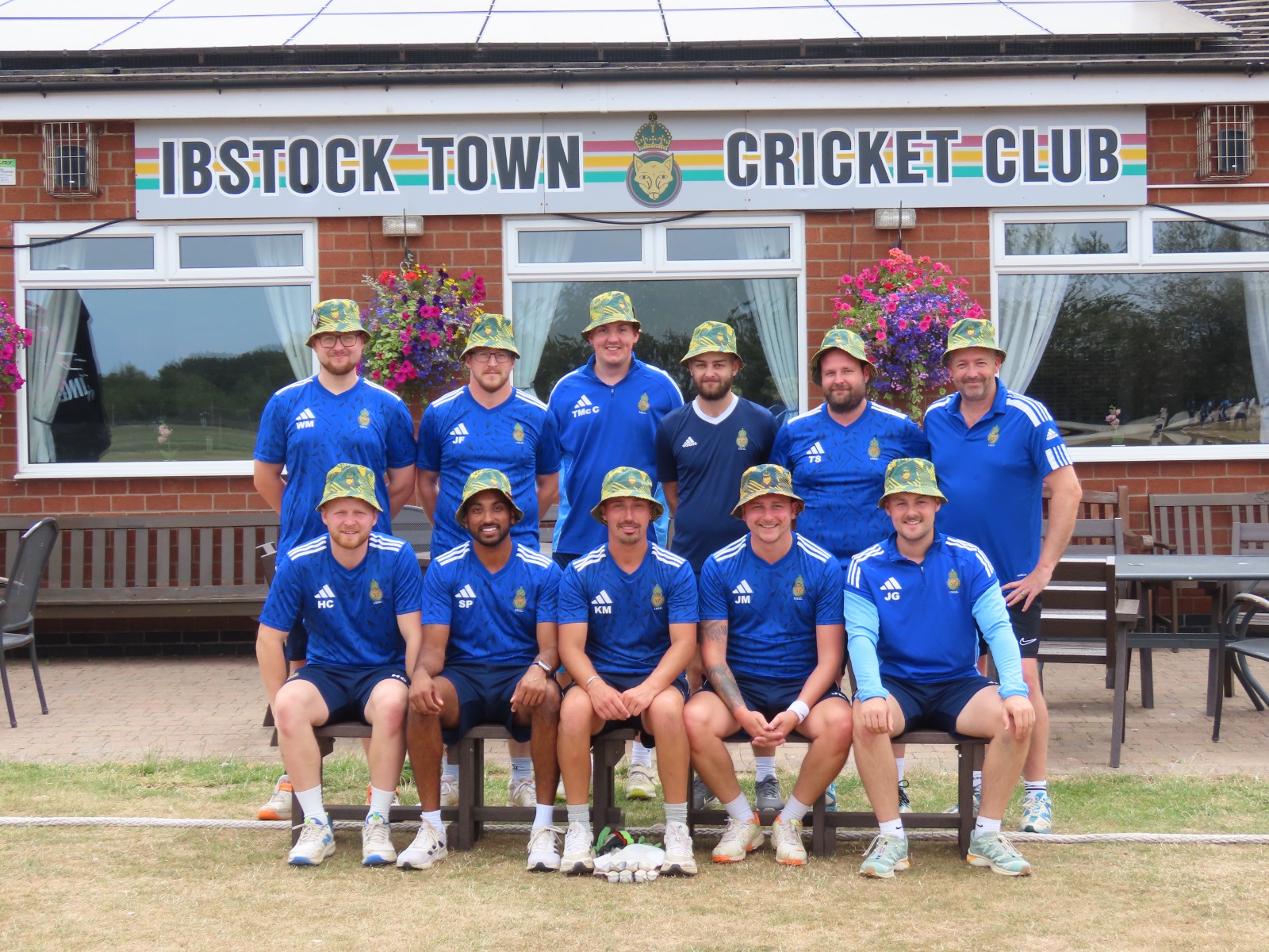 Ibstock Town CC full squad in blue kit and camo printed baggy hats in front of the clubhouse