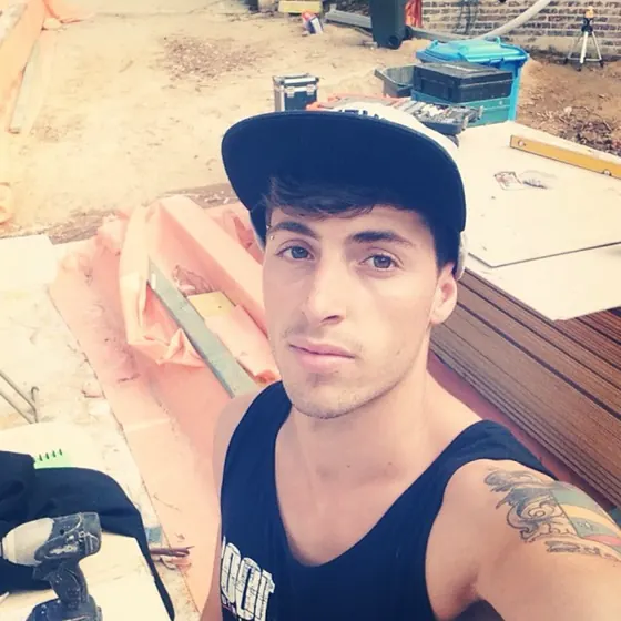 Matt Landgrebe near the end of his apprenticeship, a selfie in a cap and singlet on a building site, with a level and sheets of yellow tongue behind him.