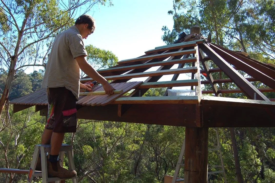 Matt Landgrebe at about 20, standing on a ladder fixing rafters on a timber gazebo roof in the bush.