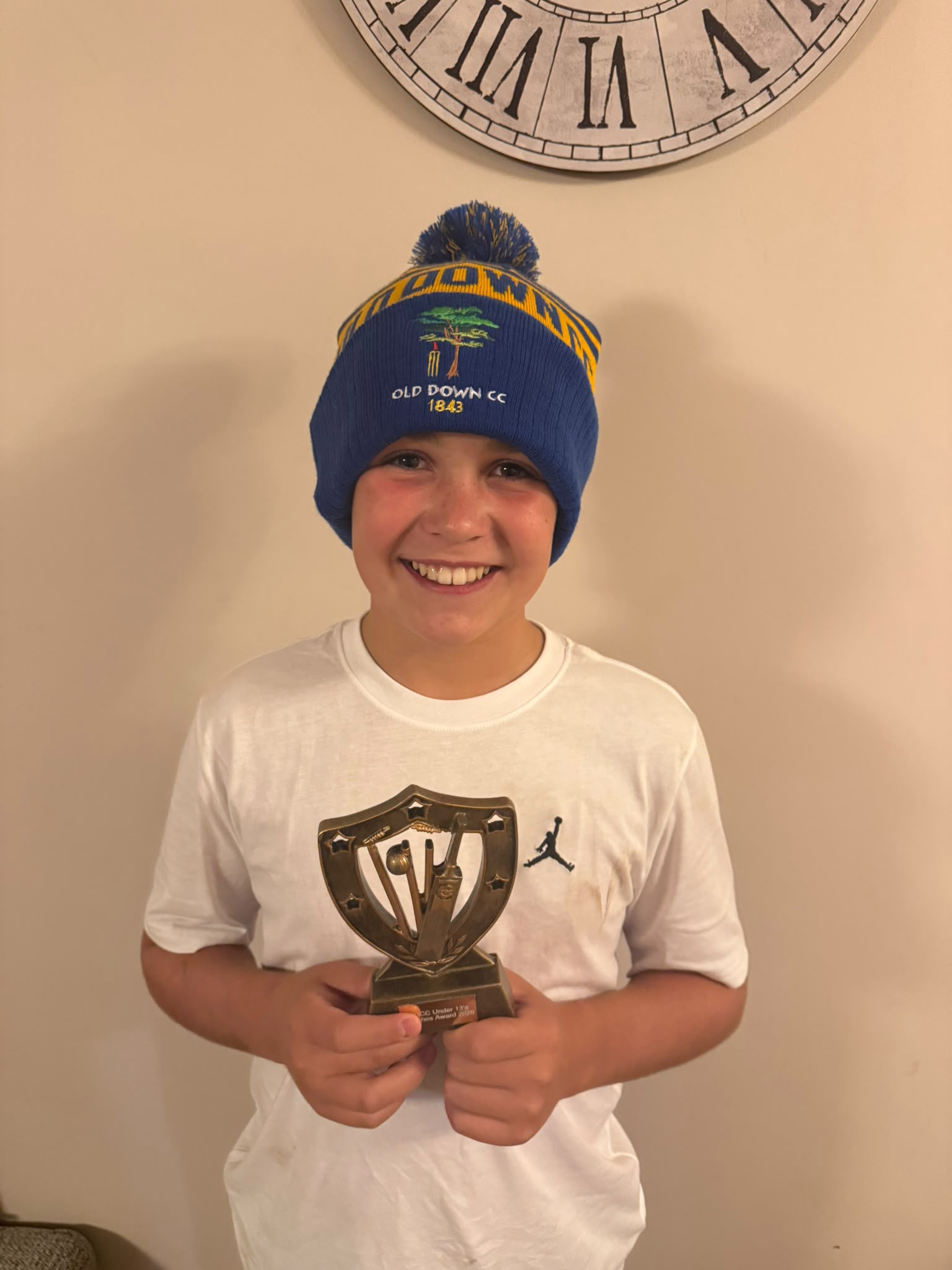 Young Old Down CC cricketer proudly holding a trophy while wearing a custom navy and yellow Old Down CC bobble hat with tree emblem and 1843 text