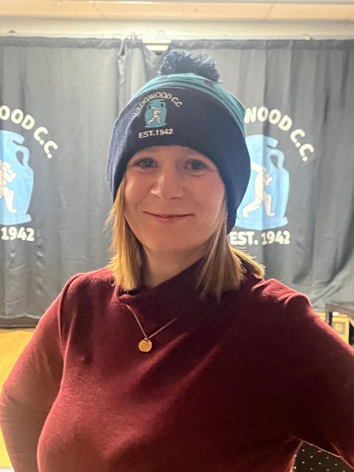 Smiling woman in a maroon top wearing a custom navy and light blue Wedgwood CC EST. 1942 bobble hat with cricketer crest, in a clubhouse with matching club banners behind
