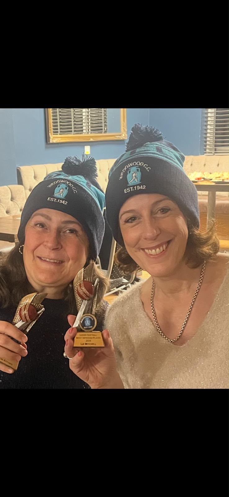 Two smiling women in matching navy and light blue Wedgwood CC EST. 1942 bobble hats holding trophies at an awards event, club banner in background