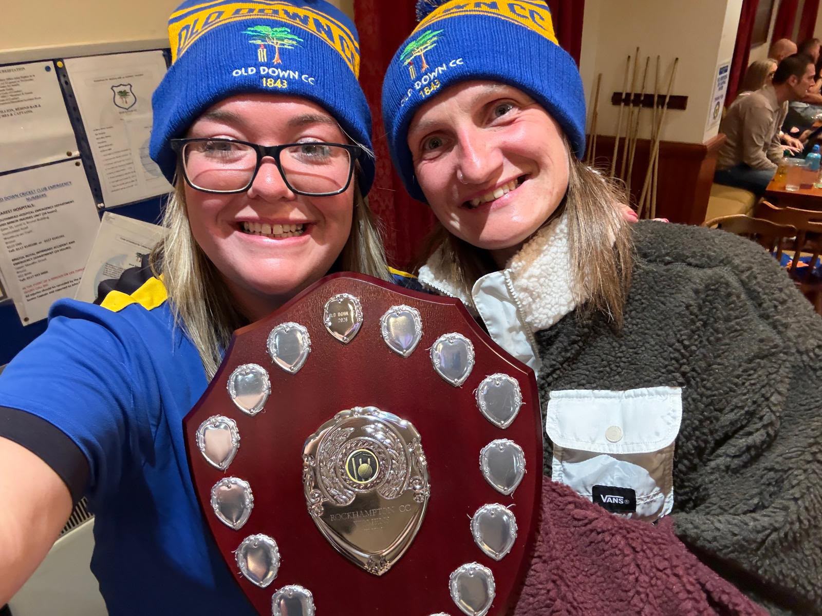 Two smiling women in matching blue and yellow Old Down CC 1843 bobble hats holding a Rockhampton CC shield at a club presentation night