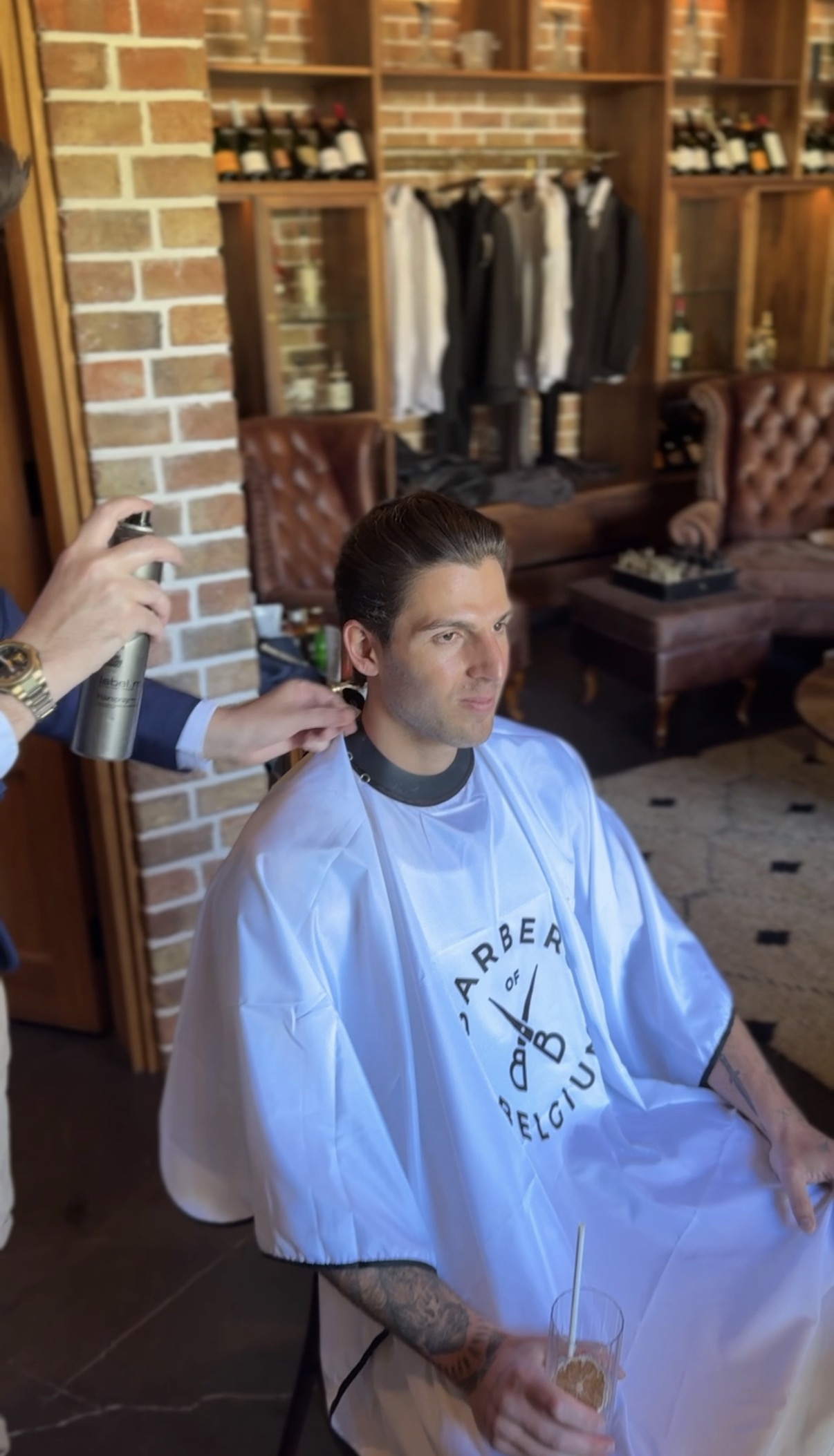 Groom being styled at a luxury wedding venue with Barbers of Belgium cape, suit racks and bar in background
